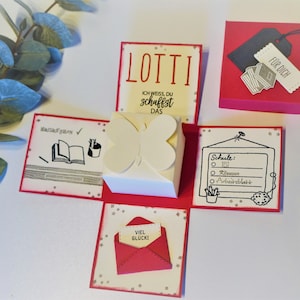 Peut inclure: Une boîte à explosion blanche avec un motif de papillon rouge. La boîte contient une enveloppe rouge avec les mots "VIEL GLÜCKI" à l'intérieur. La boîte a également une étiquette rouge avec les mots "FÜR DICH" dessus. La boîte est décorée de paillettes argentées.