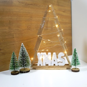 Könnte beinhalten: Weihnachtsbaumdekoration aus Holz mit dem Wort "XMAS" in weißen Buchstaben. Der Baum ist mit Lichterketten geschmückt und von kleinen, grünen Weihnachtsbäumen umgeben. Der Hintergrund ist eine Holzplatte.