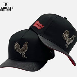 May include: Two black baseball caps with a silver rooster emblem. One cap has the text "Ganar o Ganar" in red. The caps have a black brim and a red interior. The Ferreti Premium logo is in the upper left corner.