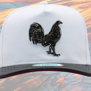 May include: A white baseball cap with a black rooster emblem on the front. The brim is dark gray with a red trim. The cap has a structured fit and a snapback closure.