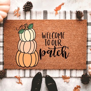 May include: A brown coir doormat with the text "Welcome to our patch" in black script. A stack of three orange pumpkins is to the left of the text.