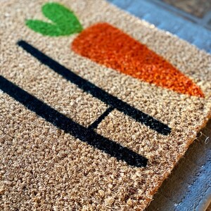 May include: A brown coir doormat with the word "Hi" in black lettering and a green and orange carrot design.