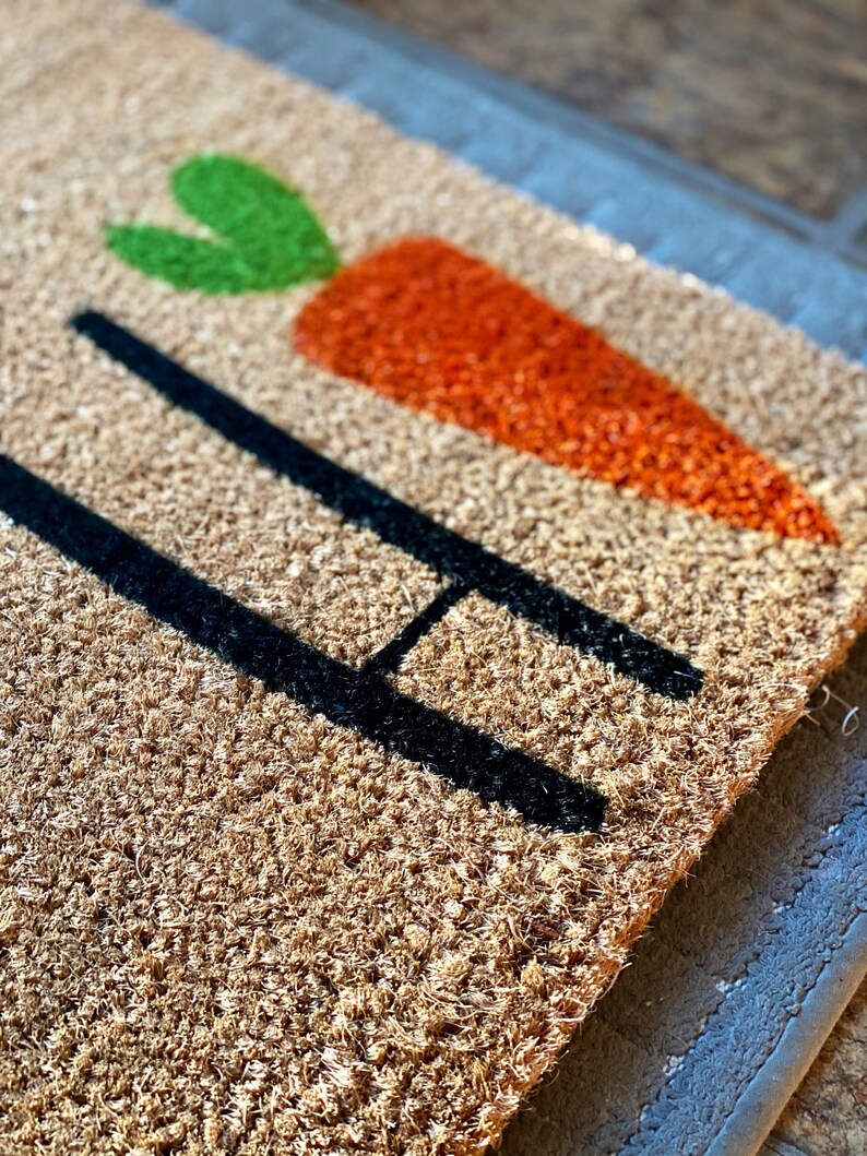 May include: A brown coir doormat with the word "Hi" in black lettering and a green and orange carrot design.