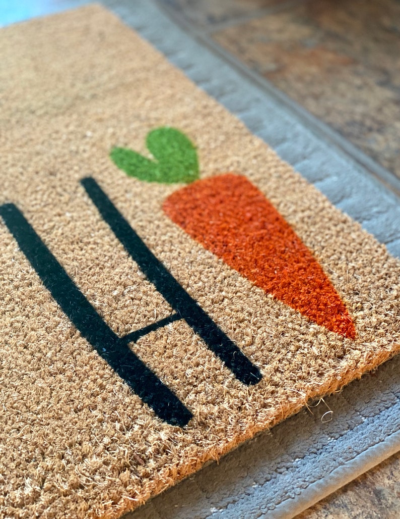 May include: A brown coir doormat with the word "Hi" in black lettering and a green and orange carrot graphic.