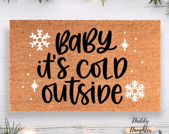 May include: A rectangular doormat with the phrase "Baby it's cold outside" in black script, accented with white snowflakes and stars. The mat has a natural, textured brown surface, perfect for a front door.