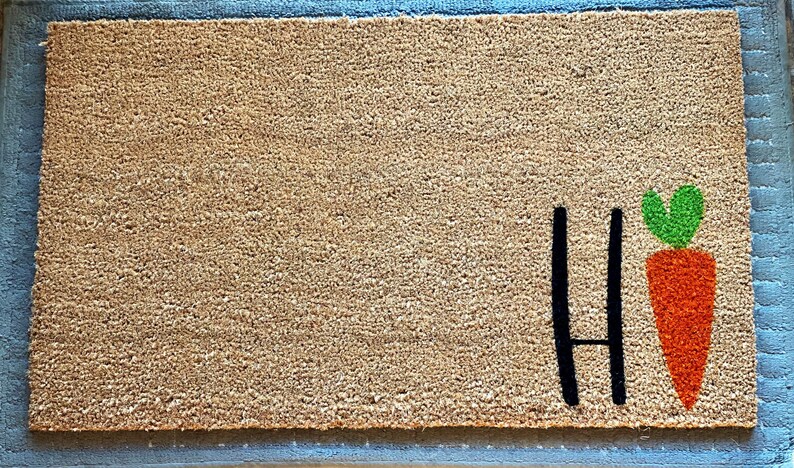 May include: A brown coir doormat with the word "Hi" in black lettering and a green-topped orange carrot graphic.