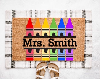 May include: A rectangular doormat with a brown coir surface features a colorful crayon design. The text "Mrs. Smith" is displayed in black. The mat is placed on a black and white checkered rug, with a pair of sandals at the bottom.