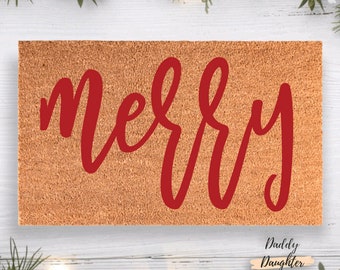 May include: A rectangular tan coir doormat featuring the word merry written in a bold red script font. The mat has a coarse, natural fiber texture and is centered against a white wooden background with green pine branches and lights.