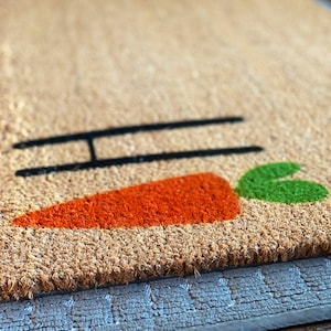 May include: A brown coir doormat with the word "Hi" and a carrot design in orange and green.
