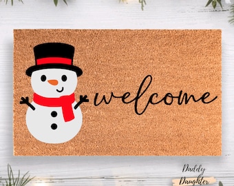 May include: A rectangular doormat with a snowman graphic and the word "welcome" in black script. The snowman has a black top hat, red scarf, and black buttons. The mat is a light brown color.