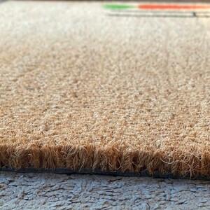 May include: Close-up of a brown coir doormat with a textured surface. The mat has a black backing.
