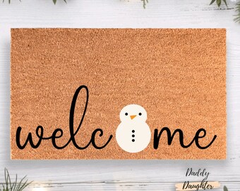 May include: A rectangular doormat with a brown coir surface. The word "welcome" is written in black cursive, with a snowman graphic replacing the "o". The snowman has a beige body, black dots for buttons, and an orange triangle nose.