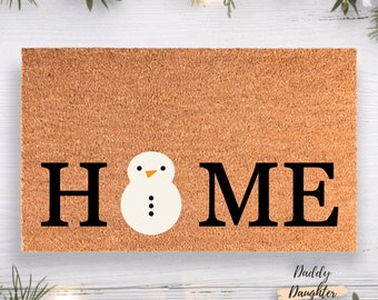 May include: A rectangular doormat with the word "HOME" in black block letters. The "O" is replaced by a snowman with a white body, black dots for buttons, and an orange beak. The mat is brown with a textured surface.