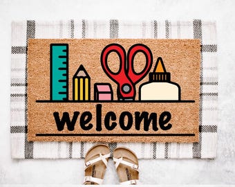 May include: A rectangular doormat with a brown coir surface features a black outline of school supplies, including a ruler, pencil, scissors, eraser, and glue bottle. The word "welcome" is written in black script. A pair of sandals is at the bottom.