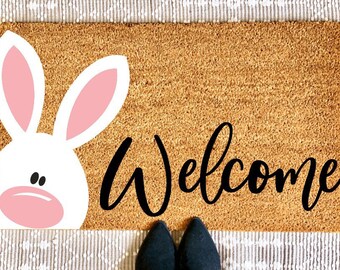 May include: A brown coir doormat with the word "Welcome" in black cursive lettering. A white bunny with pink ears and nose is partially visible on the left side of the mat.