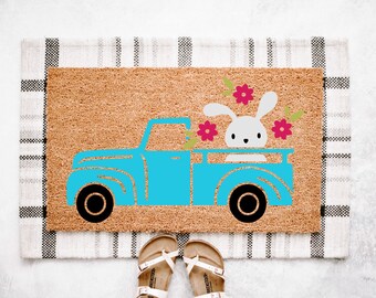 May include: A rectangular doormat with a light brown surface and a black and white plaid border. The mat features a blue truck graphic carrying a white bunny and pink flowers. Black wheels complete the truck design.