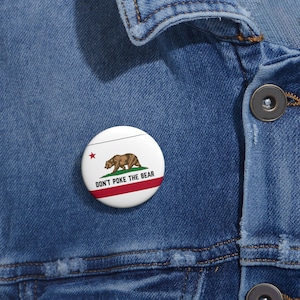 May include: A round white button with the California state flag design. The button features a brown bear, a red star, and the text "DON'T POKE THE BEAR" in black. The button is attached to a blue denim jacket.