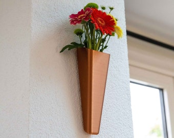 Könnte beinhalten: Eine kupferfarbene Wandvase mit dreieckiger Form, in der ein Strauß roter, rosa und gelber Blumen steckt.