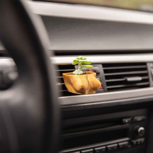 May include: A small succulent plant in a brown, 3D printed pot shaped like a bust. The pot is placed in the air vent of a car's dashboard.