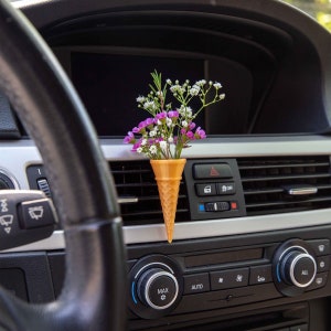 May include: A small bouquet of pink and white flowers in a cone-shaped car air freshener. The air freshener is shaped like an ice cream cone and is placed in the air vent of a car.