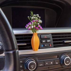 May include: A small, orange ceramic vase with a bouquet of pink and white flowers sits in the air vent of a car dashboard. The vase is attached to the vent with a clip.