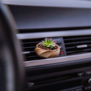Op de afbeelding: Een kleine groene succulent in een bruine pot is geplaatst in de ventilatieopening van het dashboard van een auto.
