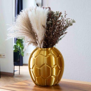 Könnte beinhalten: Eine goldfarbene Vase mit Fußballmuster, gefüllt mit getrocknetem Pampasgras und anderen getrockneten Blumen. Die Vase steht auf einem Holztisch. Die Vase hat ein einzigartiges, geometrisches Muster.