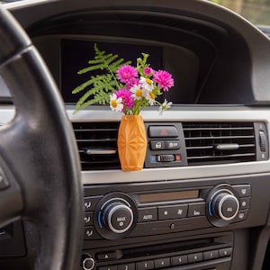 May include: A yellow geometric vase with a bouquet of pink and white flowers sits in a car's air vent. The car's dashboard is visible with controls for the radio, air conditioning, and other features.