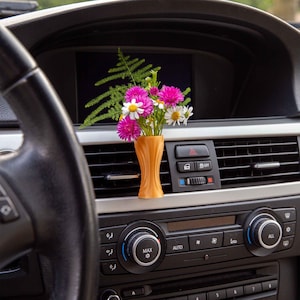 May include: A close-up of a car's interior with a vase of pink and white flowers in the center console. The vase is orange and has a modern design. The car's dashboard is black and has a variety of buttons and controls.