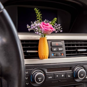 May include: A small orange vase with a textured surface holds a bouquet of pink and white flowers. The vase is placed in the air vent of a car dashboard.