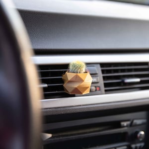 Może przedstawiać: Mały kaktus w złotej, geometrycznej doniczce, umieszczony w kratce wentylacyjnej deski rozdzielczej samochodu.