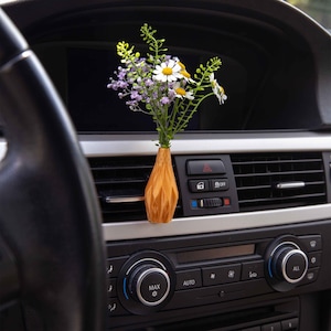 May include: A wooden vase with a geometric pattern holds a bouquet of white daisies and purple flowers in a car's air vent.