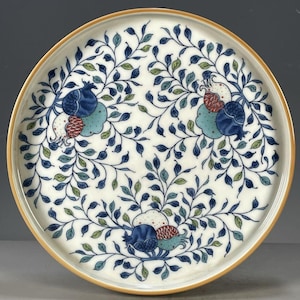 May include: Circular ceramic plate with a cream backdrop and a detailed floral pattern. The design showcases stylised blue, green, and red fruit and leaves. The plate's rim has a gold-coloured border.