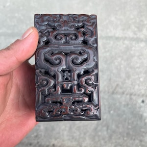 Pode incluir: Uma caixa retangular de madeira entalhada, marrom escuro, com intrincados desenhos florais e de volutas. A caixa é segurada em uma mão, mostrando as entalhes em relevo detalhadas e a rica cor da madeira. A estética geral sugere um item vintage ou antigo.