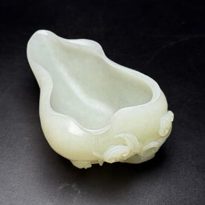 May include: A pale green, gourd-shaped bowl with a smooth, polished surface. The bowl features carved floral accents on the exterior and a wide, open top. The bowl is made of a translucent material.