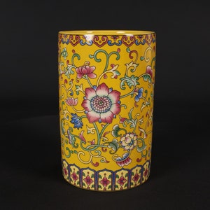 Könnte beinhalten: Eine gelbe Keramikvase mit einem Blumenmuster. Die Vase hat eine zylindrische Form und ist mit rosa, blauen und grünen Blumen und Blättern verziert. Die Vase hat einen goldenen Rand oben und unten.