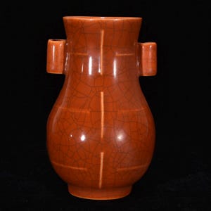 May include: A terracotta-colored ceramic vase featuring a distinct crackle glaze finish. The vessel has a wide, flared rim, a bulbous body, and two small, rectangular handles protruding from the upper sides. The surface displays a network of fine lines.