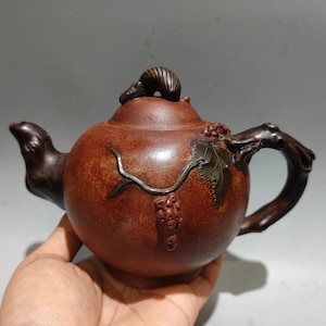 May include: A round, dark brown clay teapot featuring a textured surface with a sculpted vine branch, a single leaf, and a cluster of grapes on the side. The lid is topped with a small, curved animal figure, and the handle resembles a gnarled branch.