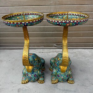 May include: A pair of decorative candle holders featuring elephant-shaped bases with teal, red, and yellow floral enamel patterns. Each elephant has a gold-toned trunk extending upward to support a wide, circular, matching enameled tray top.