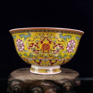 Peut inclure: Un bol en céramique jaune avec des motifs floraux et de volutes complexes en rouge, rose et vert. Le bol a un bord évasé et un pied. Le fond est noir.