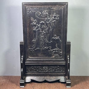 May include: A dark brown wooden table screen featuring an intricate relief carving of two figures and a crane. The rectangular panel is set within a sturdy frame with a base, showcasing detailed patterns and smooth, polished surfaces.