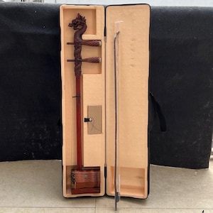 Könnte beinhalten: Eine chinesische Erhu aus Holz mit einer Drachenkopf-Schnitzerei in einem braunen Koffer mit einem schwarzen Trageriemen. Die Erhu ist ein zweisaitiges Streichinstrument.