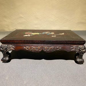 Puede incluir: Una mesa cuadrada de madera marrón oscuro con tallas ornamentadas y patas con cabeza de león. La superficie de la mesa presenta un diseño pintado con motivos florales y de aves coloridas. La mesa mide aproximadamente 46 cm de ancho y 46 cm de largo.