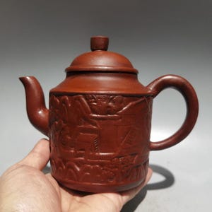 May include: A reddish-brown clay teapot featuring a cylindrical body with an embossed landscape scene depicting mountains and structures. The teapot includes a curved spout, a rounded handle, and a matching lid with a small knob handle.