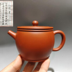 May include: A small, reddish-brown ceramic teapot with a rounded body, a curved handle, and a spout. The lid has a small knob. A handwritten note is visible in the background. The teapot is held by a hand.