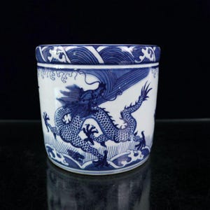 May include: A cylindrical white and blue ceramic container. The container features a detailed blue dragon design on the side, with waves at the bottom and top. The dragon is depicted with intricate scales and flowing details.