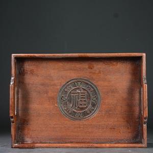 May include: A rectangular wooden tray with a carved circular design in the center. The design features a Chinese character in the center surrounded by a decorative border.