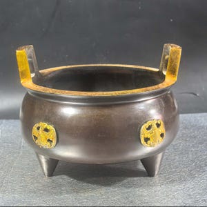 May include: A bronze-colored incense burner with three legs and two gold-colored handles. The burner has two gold-colored decorative medallions on the sides. The interior of the burner is dark, and the rim is also gold-colored.