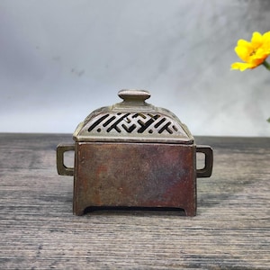 May include: An antique bronze incense burner, rectangular with a pierced, domed lid. The lid has geometric cutouts. Two small handles are on the sides. The burner sits on a wooden surface. The overall colour is a dark bronze.
