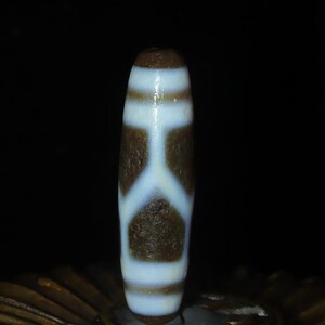 May include: A cylindrical stone bead featuring a dark brown base color with a contrasting white pattern. The design consists of horizontal bands at the top and bottom, connected by a central vertical line that branches into a Y-shape.